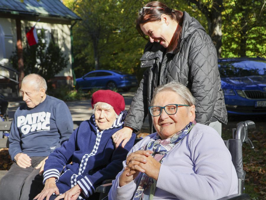 Grupa mieszkańców i opiekunka odpoczywa na ławkach w ogrodzie