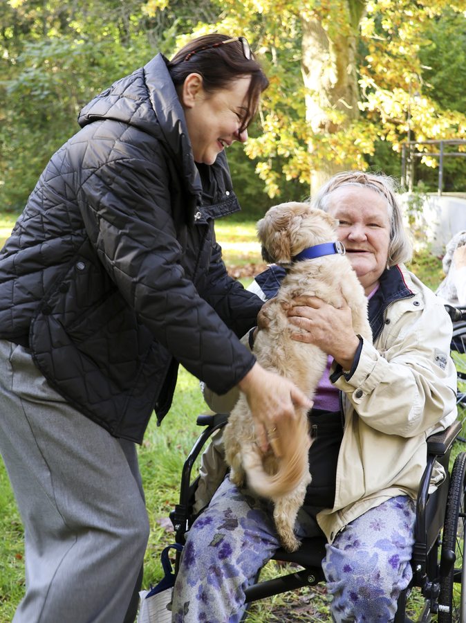 Opiekunka podaje małego psa seniorce odpoczywającej na zewnątrz